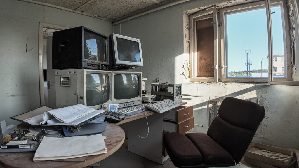 English: Interior of an abandoned room at Murru Prison with stacked vintage CRT monitors, keyboards, and office clutter on desks, showing decaying walls and a bright window looking out. Estonian: Mahajäetud ruumi interjöör Murru vanglas, kus on virnas vanad CRT-monitorid, klaviatuurid ja kontorikraam laudadel, näha on lagunevaid seinu ja hele aken, mis vaatab välja.