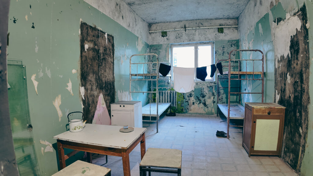English: Dilapidated room in a former Estonian prison or institutional building, showing peeling green paint on walls, two metal bunk beds, a table, and clothes drying near a bright window. Estonian: Lagunenud tuba endises Eesti vanglas või institutsionaalses hoones, kus on näha seintel kooruvat rohelist värvi, kaks metallist narivoodit, laud ja riided kuivamas heleda akna lähedal.