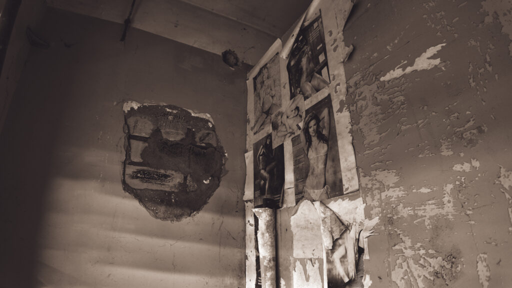 English: Dilapidated room in an abandoned Estonian building, featuring walls with peeling paint, a large hole, and old, tattered pin-up style posters. Estonian: Lagunenud tuba mahajäetud Eesti hoones, kus on seintel kooruv värv, suur auk ja vanad, rebenenud pin-up stiilis plakatid.