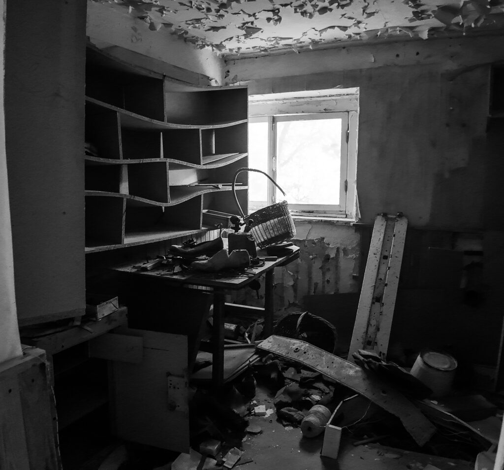 English: Black and white interior of an abandoned, decaying room at Murru Prison, showing peeling paint, empty shelves, a cluttered table, and debris on the floor, with a bright window. Estonian: Mustvalge siseruumide vaade mahajäetud, lagunevale toale Murru vanglas, kus on näha kooruvaid värve, tühje riiuleid, segamini lauda ja prügi põrandal, koos heleda aknaga.