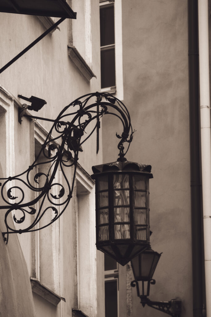 English: Close-up of an ornate, vintage wrought-iron street lamp with decorative scrolls, mounted on a historical building facade in Tallinn Old Town. Estonian: Lähivõte ornamendiga, vanaaegsest sepistatud tänavavalgustist dekoratiivsete keeristega, mis on kinnitatud ajaloolise hoone fassaadile Tallinna vanalinnas.