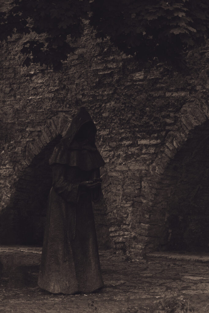 English: Black and white close-up of a dark, hooded, faceless monk statue with clasped hands, set against a tiled roof in Tallinn Old Town. Estonian: Mustvalge lähivõte tumedast, kapuutsiga, näota munga kujust, mille käed on palveasendis, Tallinna vanalinna katuse taustal.