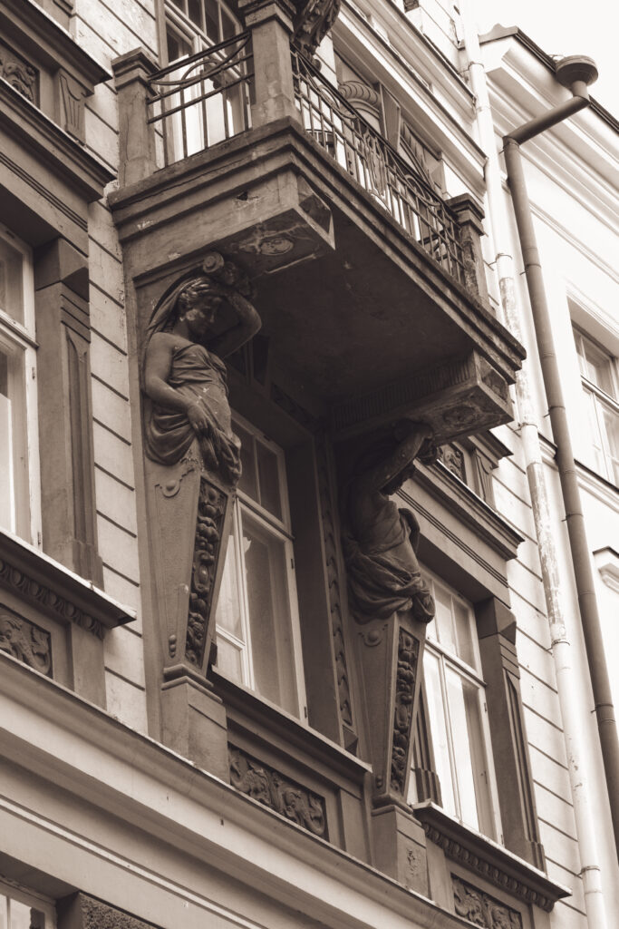 English: Close-up of a historical building facade in Tallinn Old Town, featuring an ornate balcony supported by classical sculpted figures. Estonian: Lähivõte ajaloolise hoone fassaadist Tallinna vanalinnas, millel on ornamendiga rõdu, mida toetavad klassikalised skulptuurfiguurid