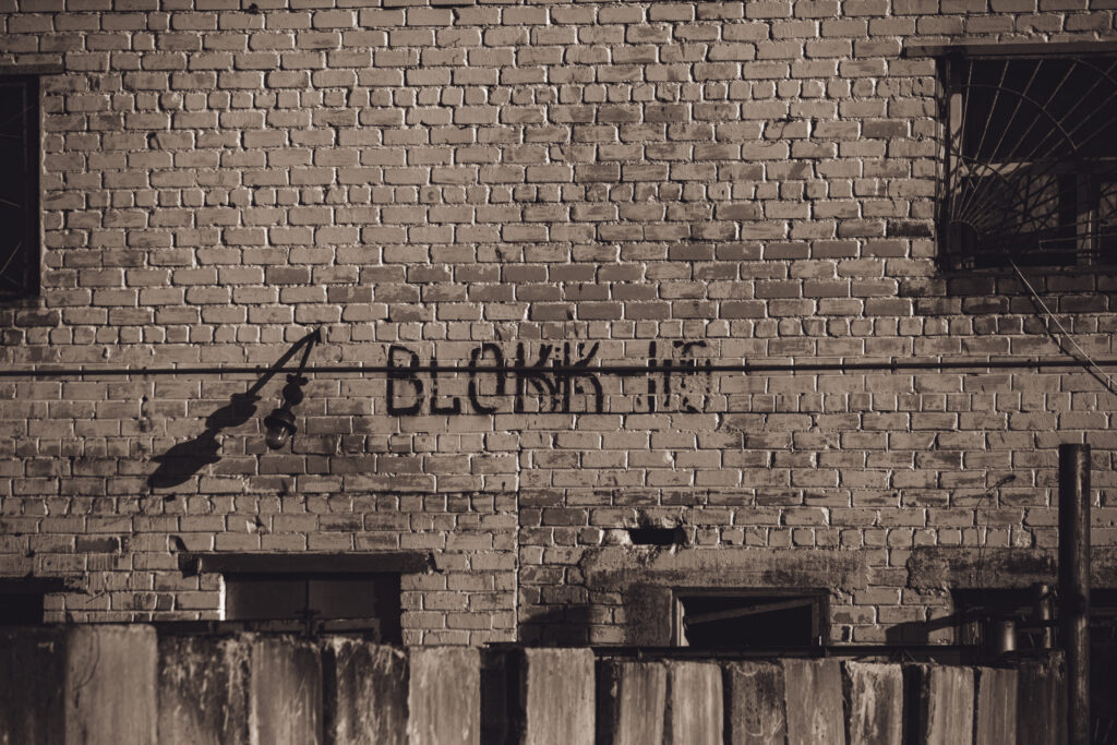 English: A weathered light-colored brick wall at Murru Prison with dark graffiti reading "BLOKK 14" and windows, one with bars, creating a grim atmosphere. Estonian: Ilmastikust kulunud heledat värvi telliskivisein Murru vanglas, millel on tume grafiti "BLOKK 14" ja aknad, millest üks on trellidega, luues sünge atmosfääri.