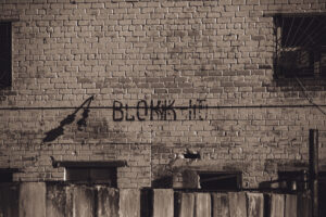 English: A weathered light-colored brick wall at Murru Prison with dark graffiti reading "BLOKK 14" and windows, one with bars, creating a grim atmosphere. Estonian: Ilmastikust kulunud heledat värvi telliskivisein Murru vanglas, millel on tume grafiti "BLOKK 14" ja aknad, millest üks on trellidega, luues sünge atmosfääri.