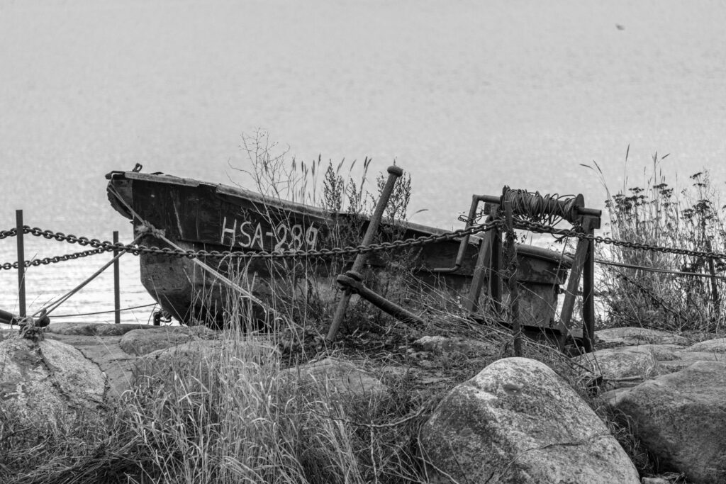 Old wooden boat marked HSA-289 behind chains and a rusty winch on a rocky shore, black and white