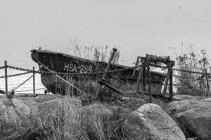 Old wooden boat marked HSA-289 behind chains and a rusty winch on a rocky shore, black and white