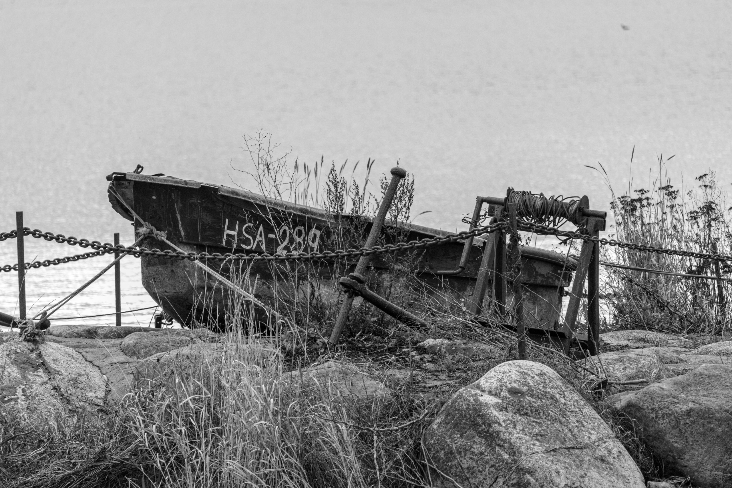 Old wooden boat marked HSA-289 behind chains and a rusty winch on a rocky shore, black and white