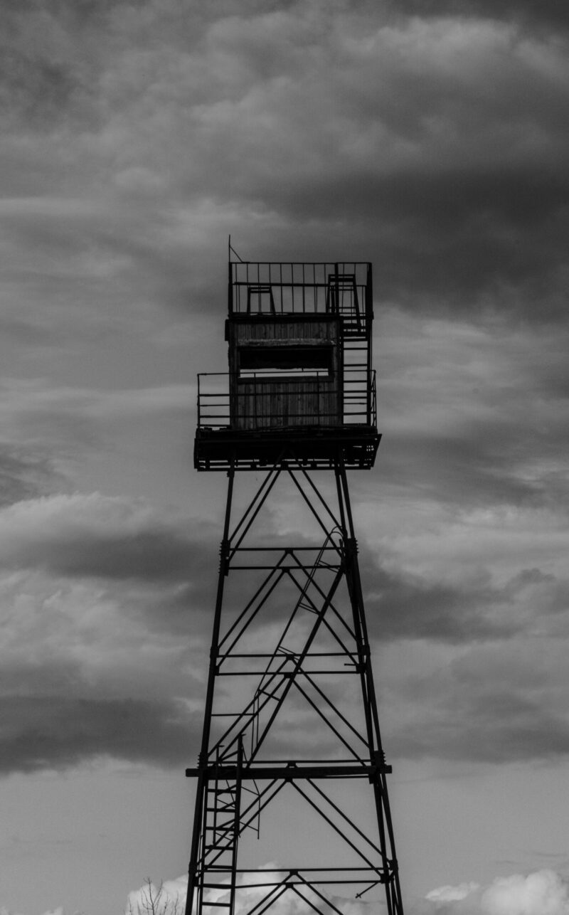 A derelict watchtower standing in a decaying building, captured amidst overgrown vegetation and worn-out structures / Hõõguv vaatetorn lagunevas hoones, jäädvustatud üle kasvanud taimestiku ja kulunud struktuuride keskel