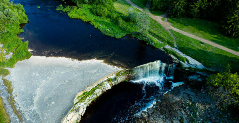 English: Aerial view of the wide Jägala Waterfall in Estonia, showing the river flowing over the natural limestone formation into turbulent water below, surrounded by lush green banks. Estonian: Droonivaade laiale Jägala joale Eestis, kus jõgi voolab üle loodusliku paekivist moodustise allolevaks tormiseks veeks, ümbritsetud lopsakate roheliste kallastega.
