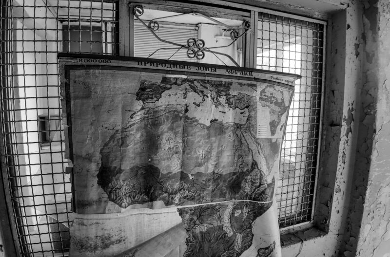 English: Black and white image of an old, tattered wall map of Africa with Cyrillic text, covering a decaying window with metal bars inside Murru Prison, Estonia. Estonian: Mustvalge pilt vanast, narmendavast Aafrika seinakaardist kirillitsas tekstiga, mis katab lagunevat, metallvarbade akent Murru vangla sees, Eestis.