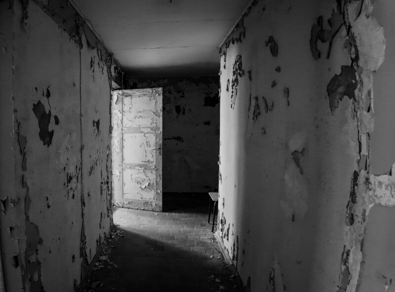 English: Black and white image of a long, decaying corridor with extensive peeling paint on walls and an open doorway leading to a dark room, inside an abandoned building in Estonia. Estonian: Mustvalge pilt pikast, lagunevast koridorist, kus seintel on ulatuslikult kooruv värv ja avatud ukseava viib pimedasse ruumi, mahajäetud hoones Eestis.