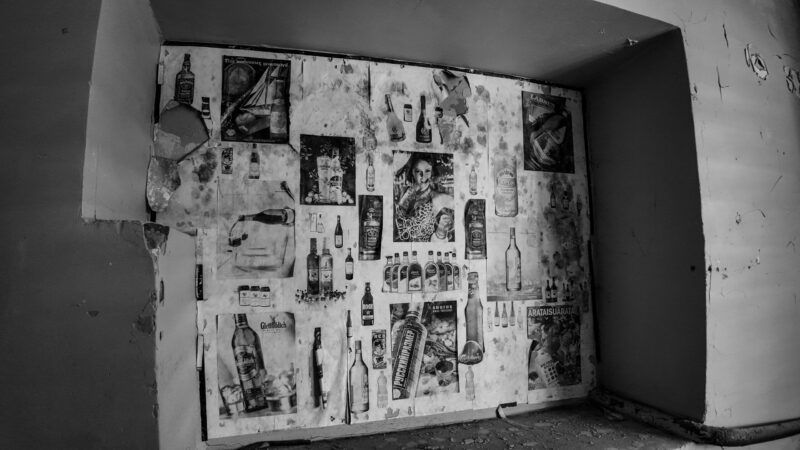 English: Black and white image of a decaying wall in Murru Prison covered with old, peeling advertisements for various alcoholic beverages, revealing crumbling plaster underneath. Estonian: Mustvalge pilt Murru vangla lagunevast seinast, mis on kaetud vanade, kooruvate alkoholireklaamidega, paljastades murenevat krohvi.