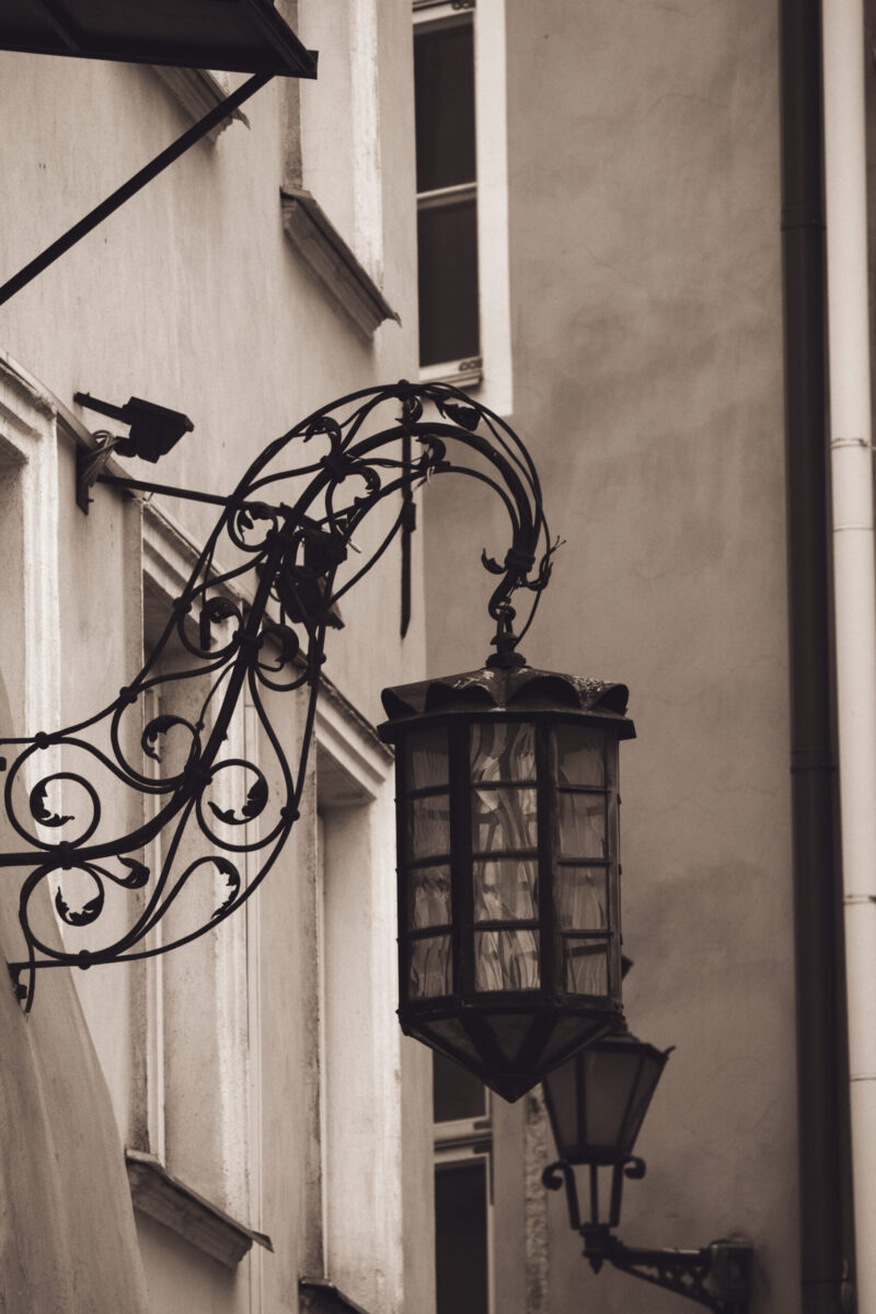 English: Close-up of an ornate, vintage wrought-iron street lamp with decorative scrolls, mounted on a historical building facade in Tallinn Old Town. Estonian: Lähivõte ornamendiga, vanaaegsest sepistatud tänavavalgustist dekoratiivsete keeristega, mis on kinnitatud ajaloolise hoone fassaadile Tallinna vanalinnas.
