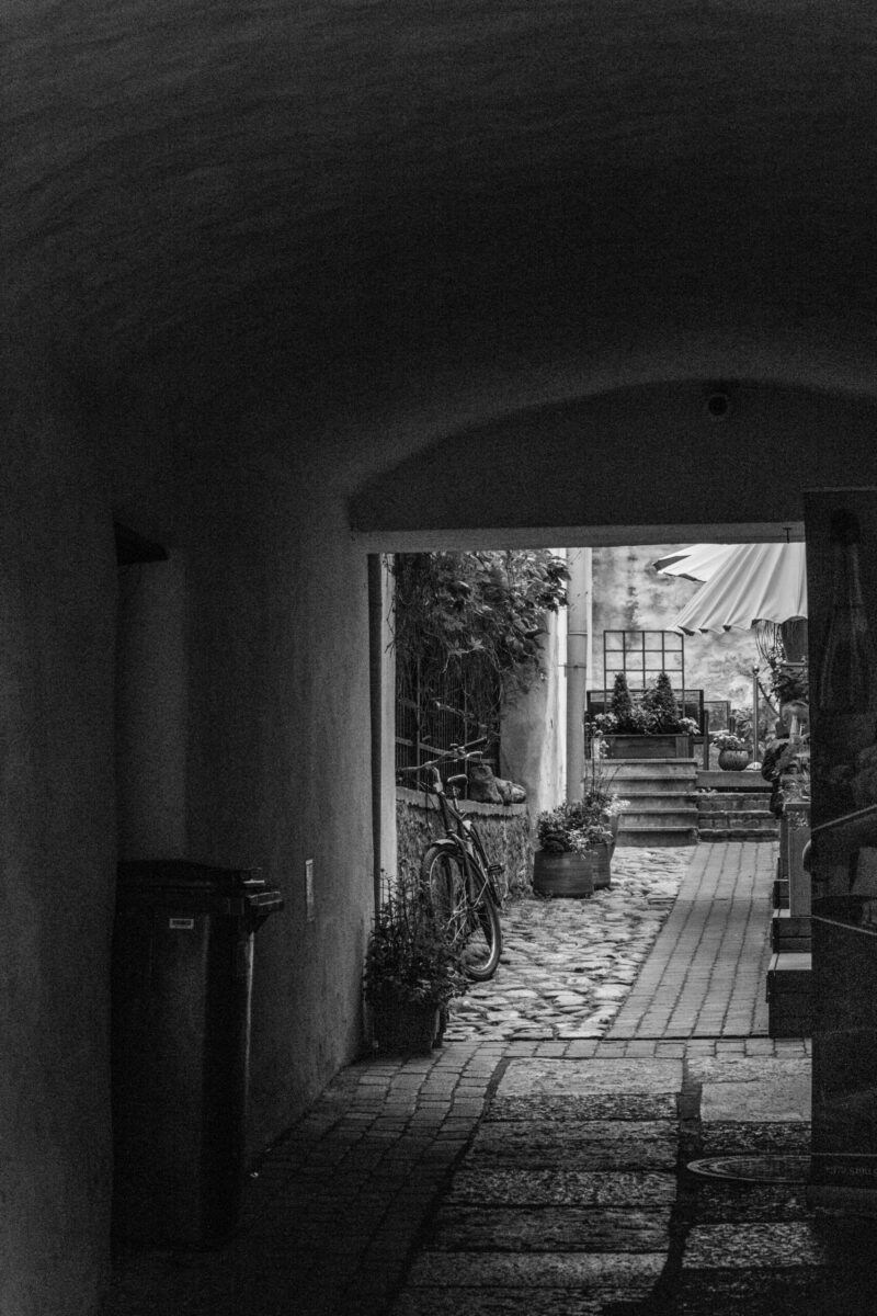 English: Black and white view from a dark arched passage into a bright cobblestone alleyway in Tallinn Old Town, with a bicycle and potted plants. Estonian: Mustvalge vaade pimedast kaarvõlviga käigust valgusküllasele munakividega kaetud alleele Tallinna vanalinnas, jalgratta ja potitaimedega