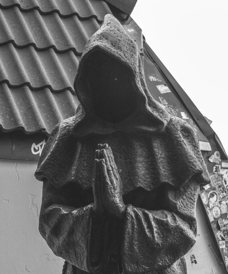 English: Black and white close-up of a dark, hooded, faceless monk statue with clasped hands, set against a tiled roof in Tallinn Old Town. Estonian: Mustvalge lähivõte tumedast, kapuutsiga, näota munga kujust, mille käed on palveasendis, Tallinna vanalinna katuse taustal.