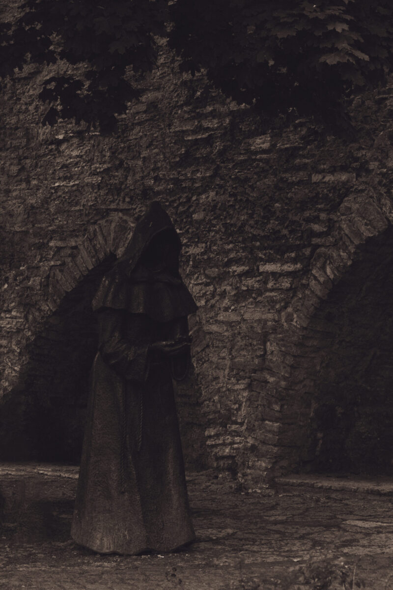 English: Black and white close-up of a dark, hooded, faceless monk statue with clasped hands, set against a tiled roof in Tallinn Old Town. Estonian: Mustvalge lähivõte tumedast, kapuutsiga, näota munga kujust, mille käed on palveasendis, Tallinna vanalinna katuse taustal.