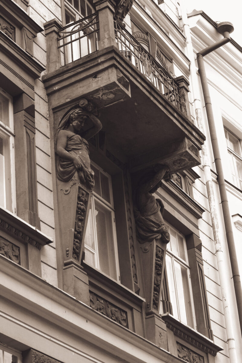 English: Close-up of a historical building facade in Tallinn Old Town, featuring an ornate balcony supported by classical sculpted figures. Estonian: Lähivõte ajaloolise hoone fassaadist Tallinna vanalinnas, millel on ornamendiga rõdu, mida toetavad klassikalised skulptuurfiguurid