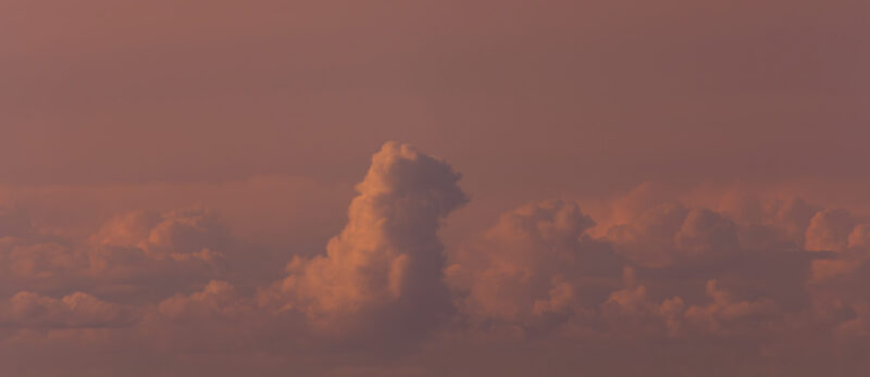 English: Wide shot of a sky at sunset or sunrise, featuring warm orange and pink-toned clouds, with one prominent cumulus formation. Estonian: Lai kaader päikeseloojangu- või päikesetõusuajast, kus on näha soojades oranžides ja roosades toonides pilvi, mille seas on üks silmapaistev rünkpilve formatsioon
