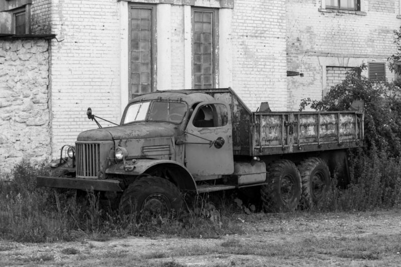 English: Black and white image of an old, rusted ZIL-157 Soviet-era truck, overgrown with weeds, parked in front of a brick building at Murru Prison, Estonia. Estonian: Mustvalge pilt vanast, roostetanud Nõukogude-aegsest ZIL-157 veokist, mis on umbrohuga kaetud ja pargitud telliskivihoone ette Murru vanglas, Eestis.